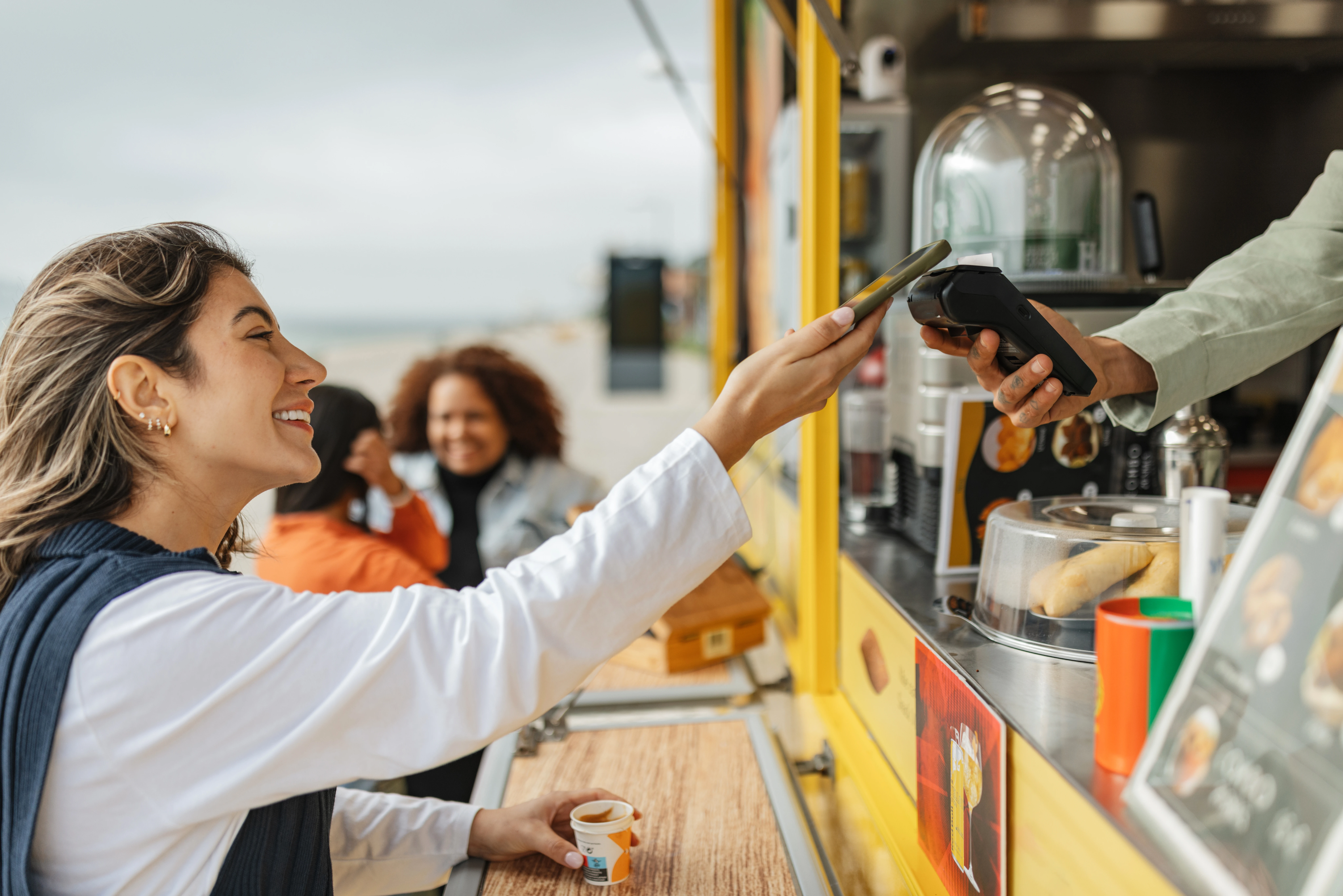 Lächelnde Kundin hält ihr Handy an ein mobiles Kartenterminal, das ihr aus einem Streetfood-Wagen entgegengehalten wird
