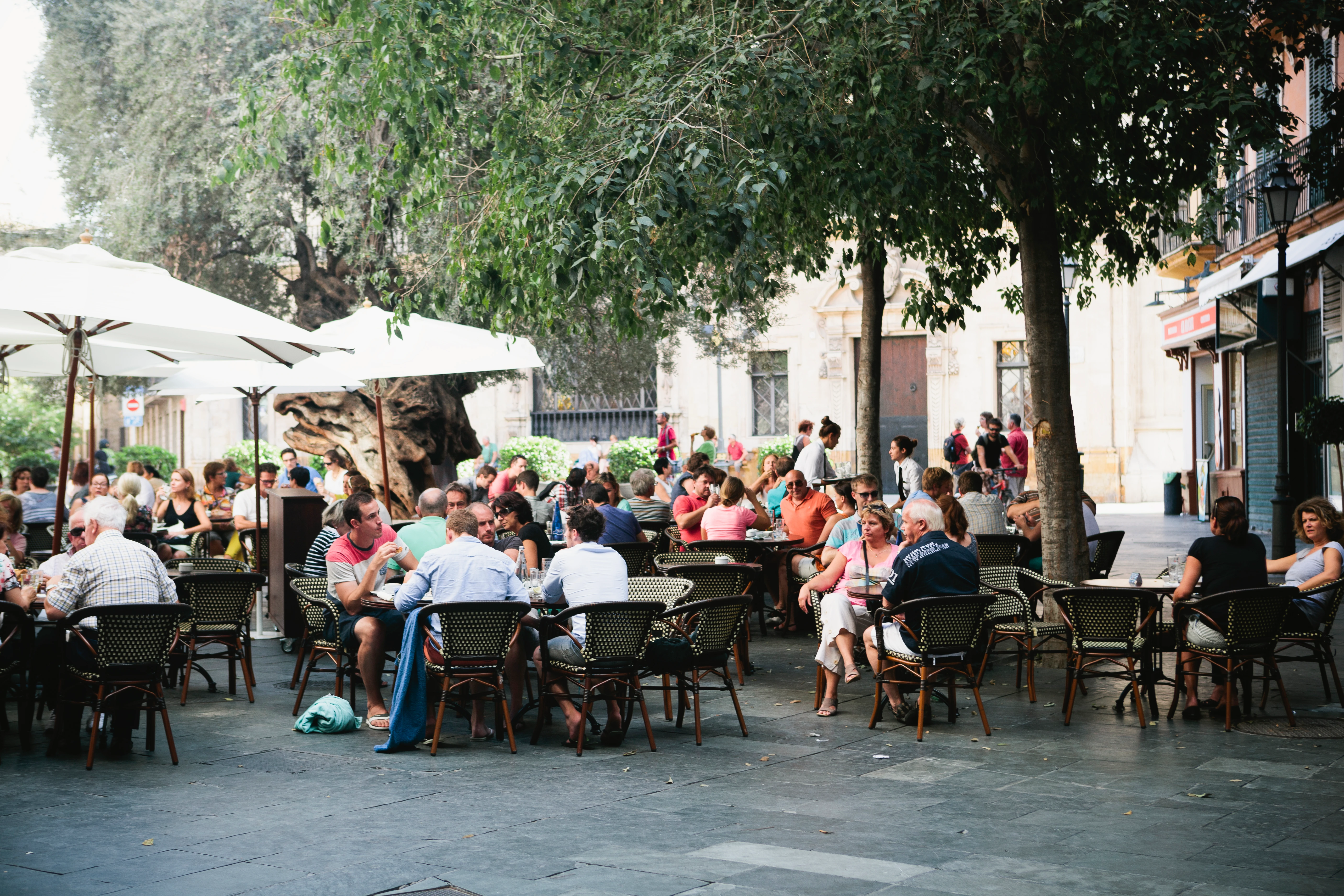 Gut besuchte Café-Terrasse mit Gästen an Tischen unter Sonnenschirmen und Bäumen auf einem Platz.