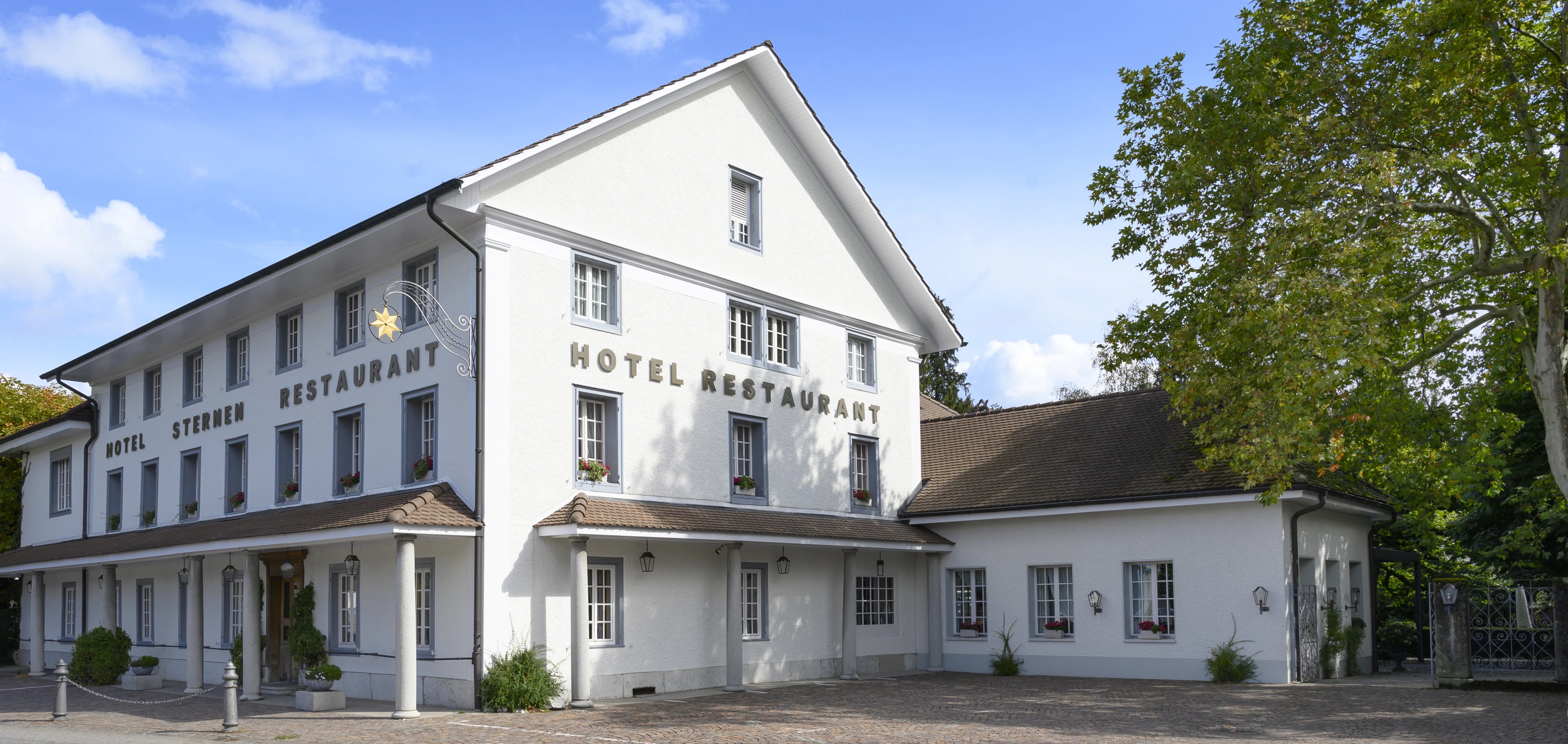 Vista esterna dell'Hotel-Ristorante Sternen: un edificio bianco con mattoni scuri, circondato da alberi e cespugli verdi.