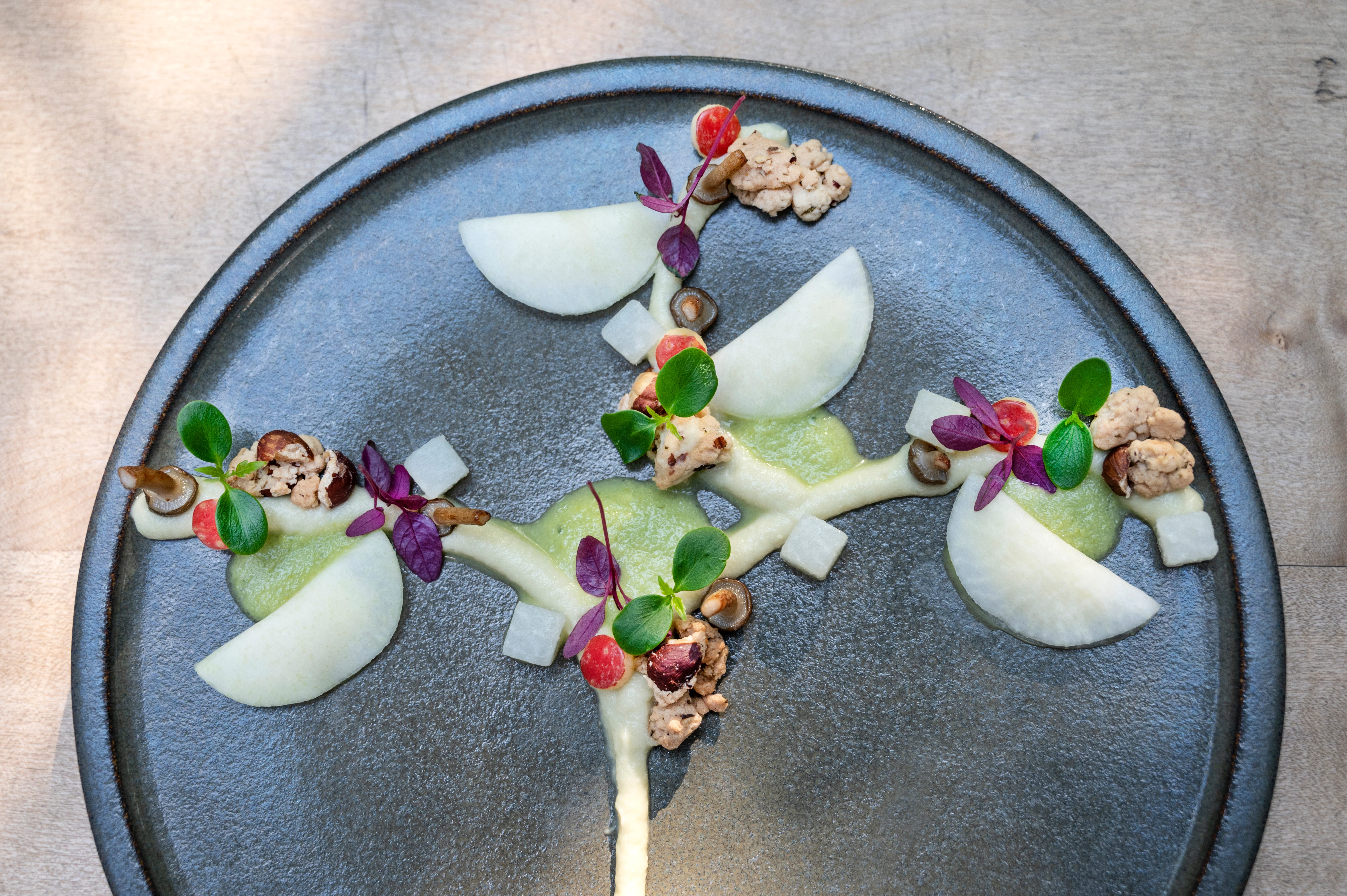 Dessert presentato con cura, con pera, crema, frutti di bosco e noci su un piatto scuro.