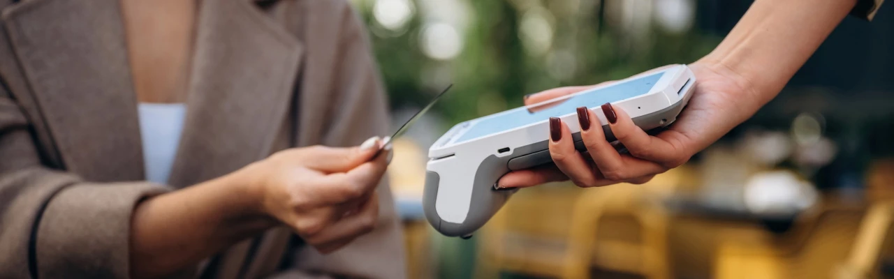 une cliente présente sa carte devant un lecteur de carte blanc permettant le paiement sans contact, à l'extérieur d'un café.